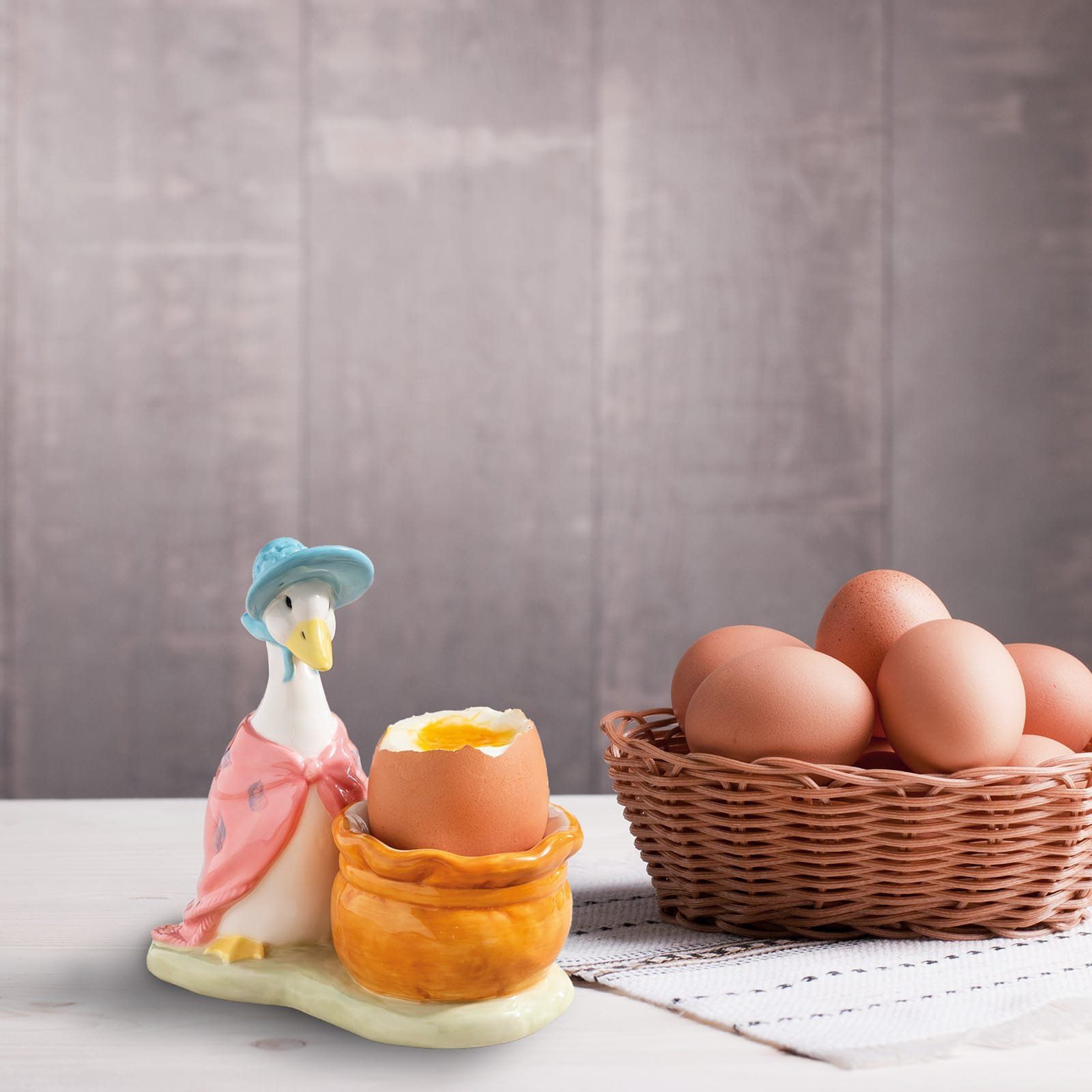 Een eierdopje in de vorm van een gans bevat een zachtgekookt ei. Een mandje met eieren en een dessin-achtig doek op een houten ondergrond.