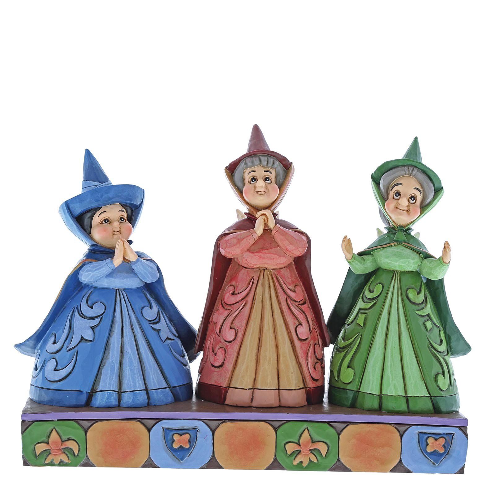 Drie Disney-feeën in kleurrijke gewaden met gevouwen handen, staande op een versierde sokkel.