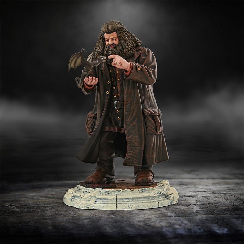 Standbeeld van Rubeus Hagrid, met een kleine draak in zijn armen, gekleed in een bruine jas, staand op een stenen sokkel.