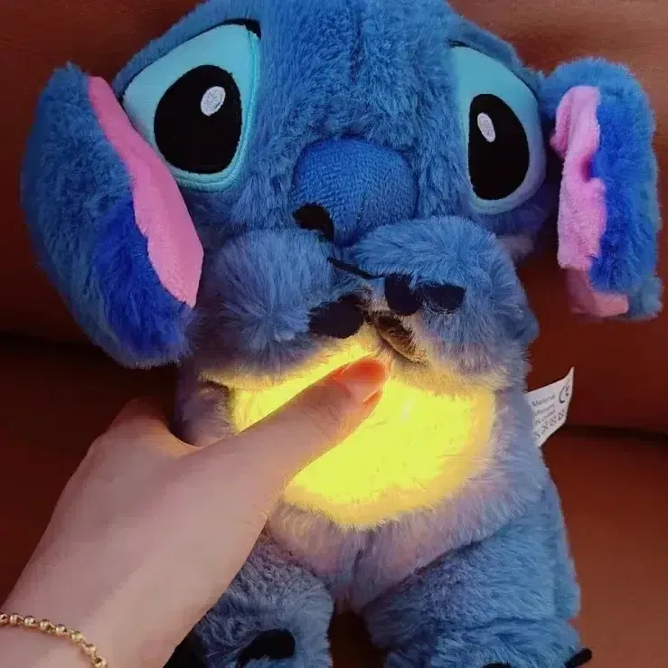 Blauwe Stitch pluche knuffel met een oplichtend geel middenstuk, vastgehouden door een hand met een gouden armband.