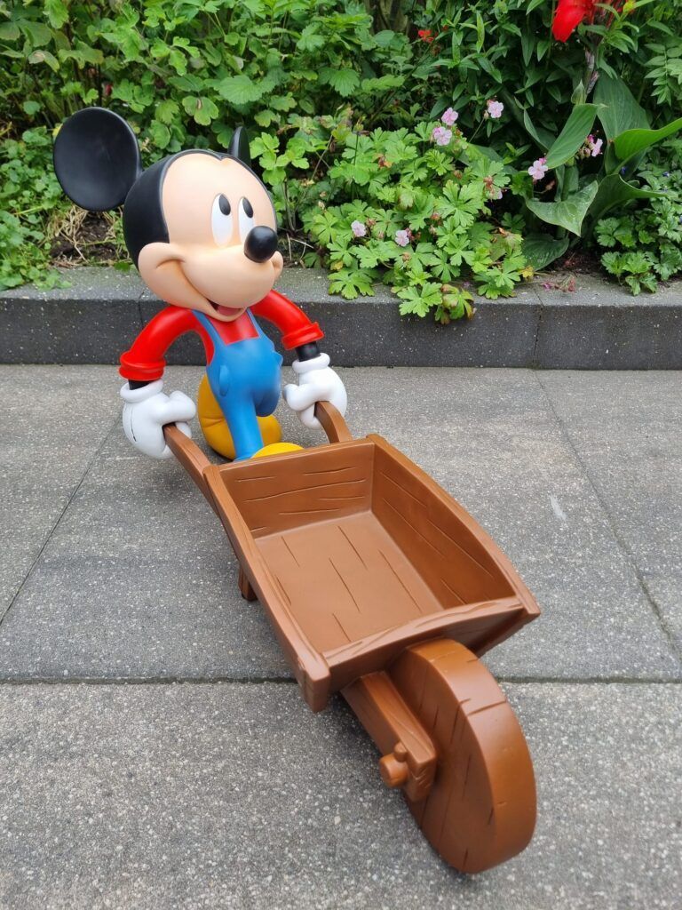 Een Mickey Mouse-figuurtje duwt een bruine kruiwagen; buiten, met een groene struik op de achtergrond.