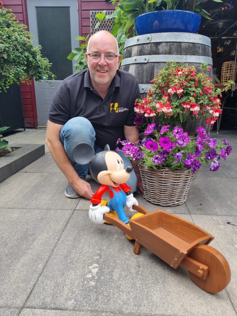 Een man knielt en poseert naast een kruiwagen met Mickey Mouse-print, bloemen in een rieten pot en een ton op de achtergrond.