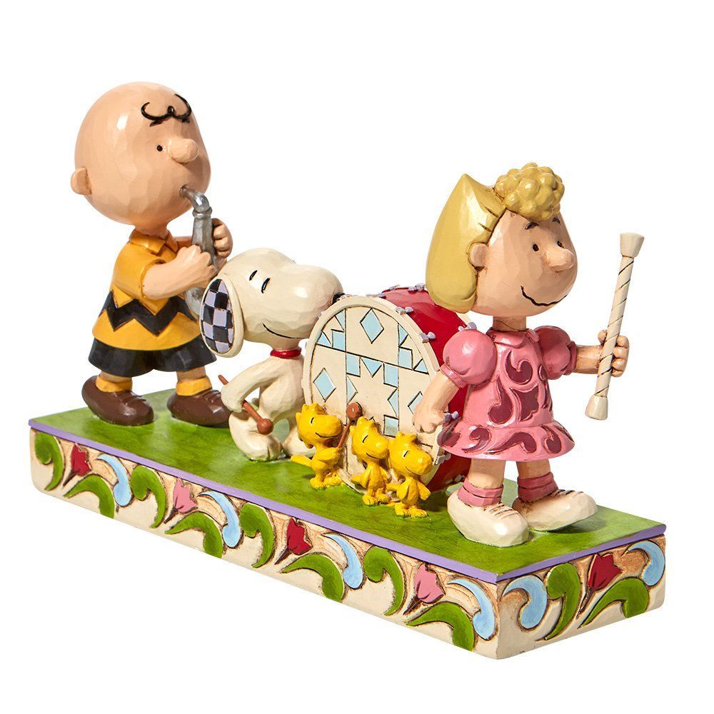 Charlie Brown die fluit speelt, Snoopy met een trommel, Sally met een baton, op een decoratieve sokkel.