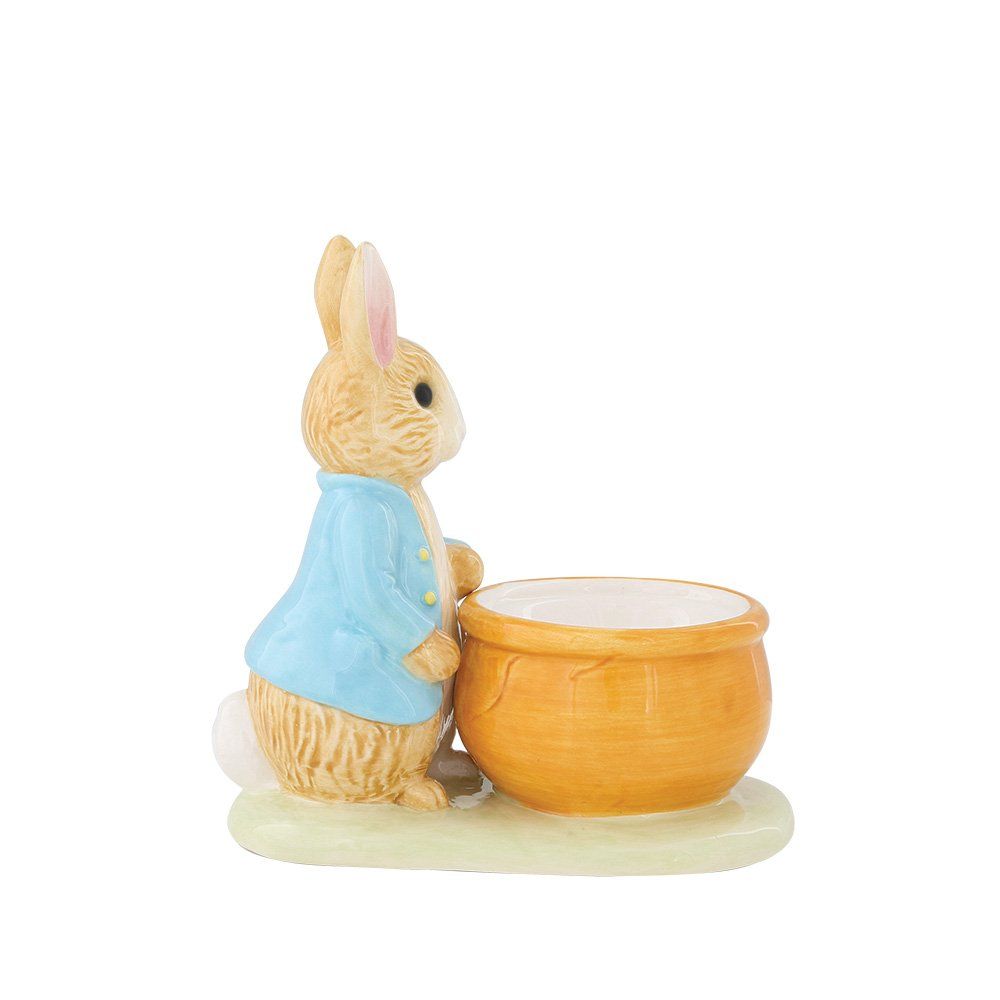 Een Peter Rabbit-beeldje dat tegen een houten kom leunt, op een grasachtige ondergrond.