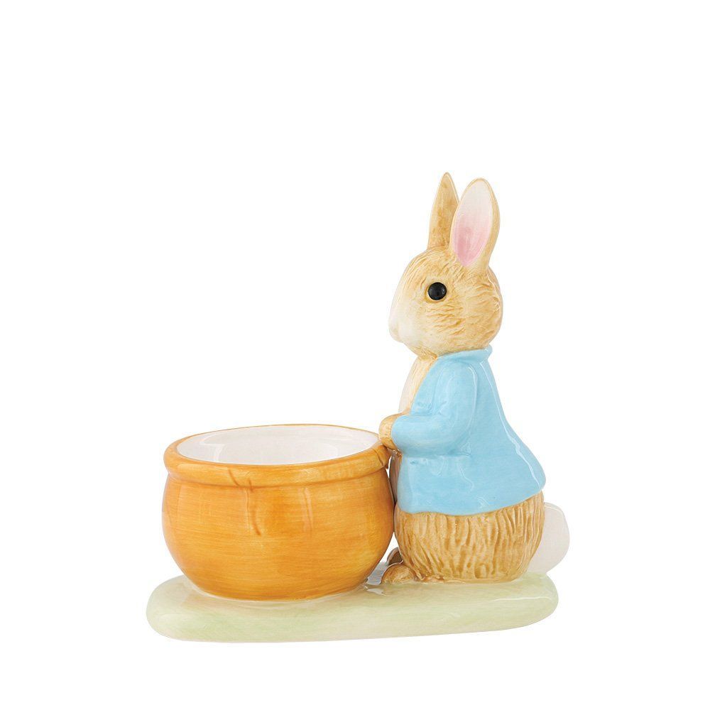 Een Peter Rabbit-beeldje dat tegen een houten kom leunt, op een grasachtige ondergrond.