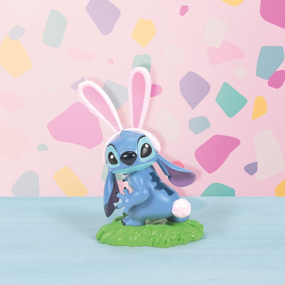Een Stitch-figuurtje met konijnenoortjes en een pluizige staart zit op groen gras. Tegen een pastelkleurige, gespikkelde achtergrond.