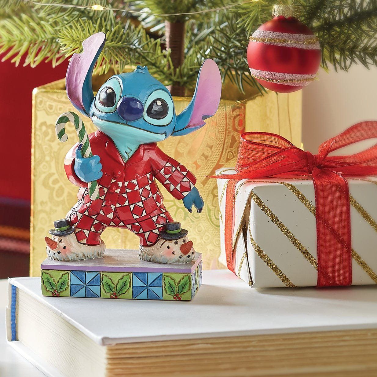 Een Stitch-figuurtje in pyjama, met een zuurstok in zijn hand, voor een kerstboom met cadeautjes.