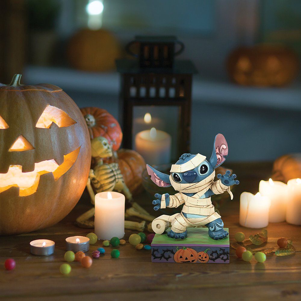 Een Stitch-figuur verkleed als mummie met pompoenen op een sokkel met Halloween-thema.