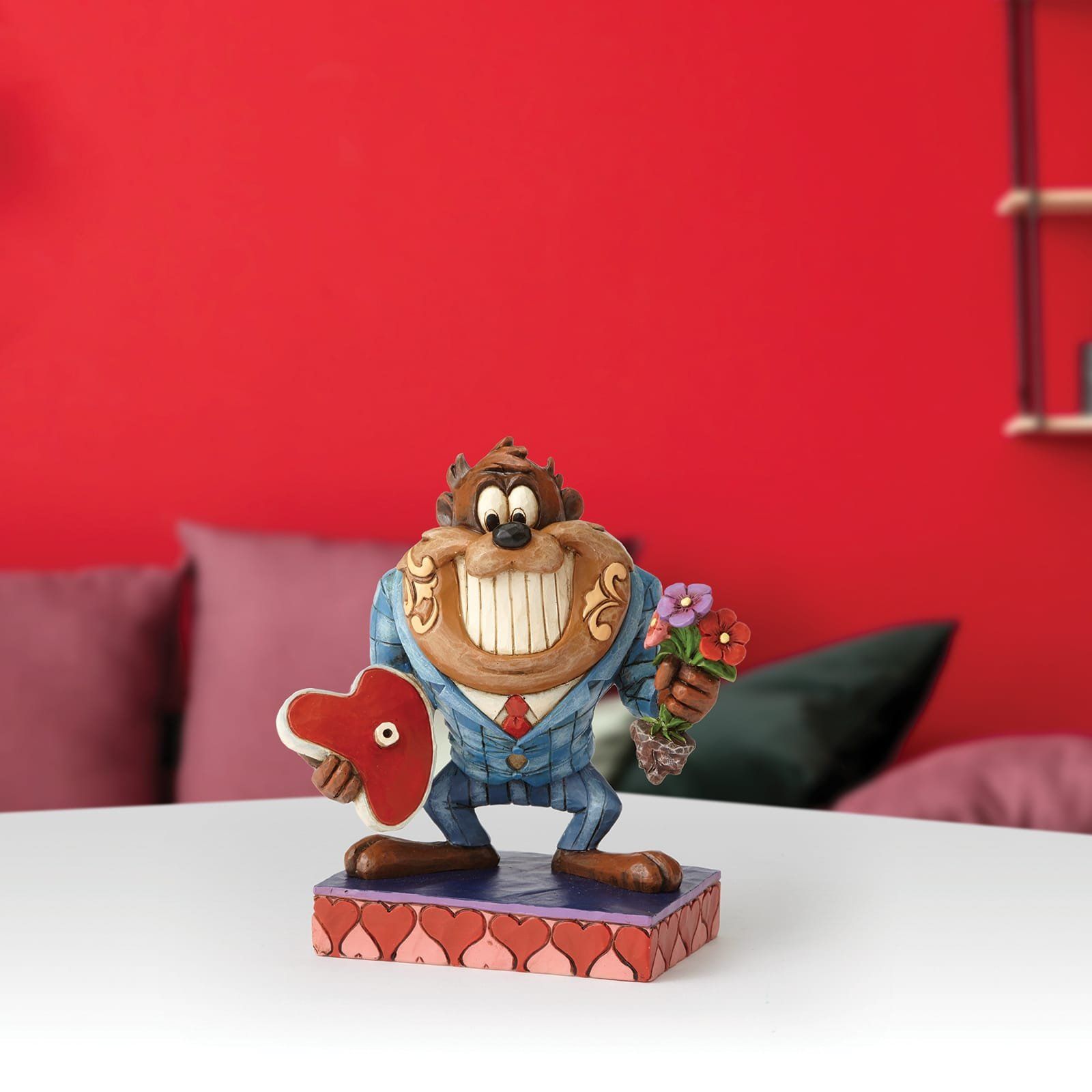 Een figuur van Taz, gekleed in een blauw pak en met een hart en bloemen in haar handen, staat tegen een rode muur.