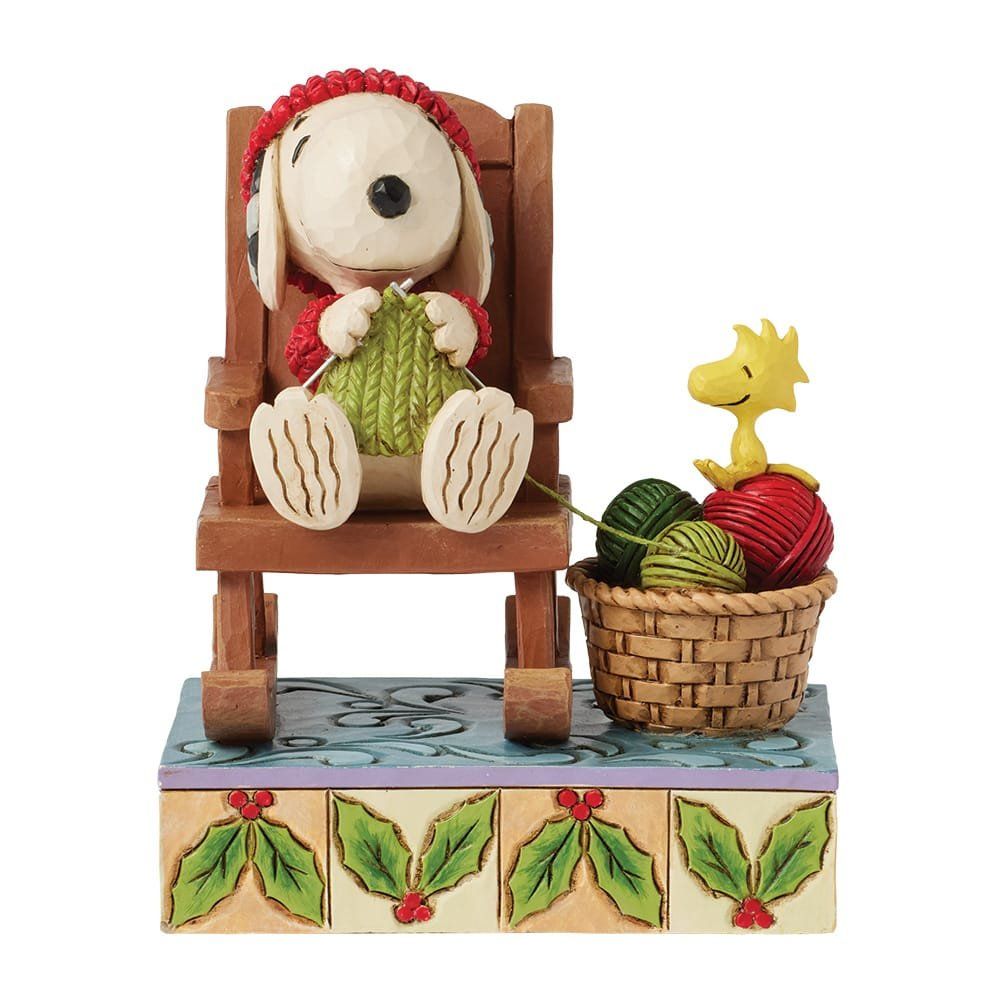 Snoopy zit te breien, met een rode muts op, in een schommelstoel, met Woodstock en garen in een mandje.