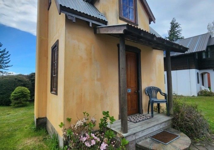 Small, yellow house with a porch. Brown door, window, and roof. A chair and some flowers are visible.