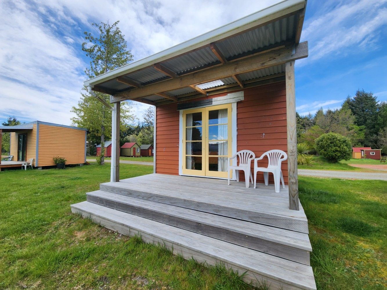 Red cabin with wooden porch and two white chairs on green grass under blue sky.