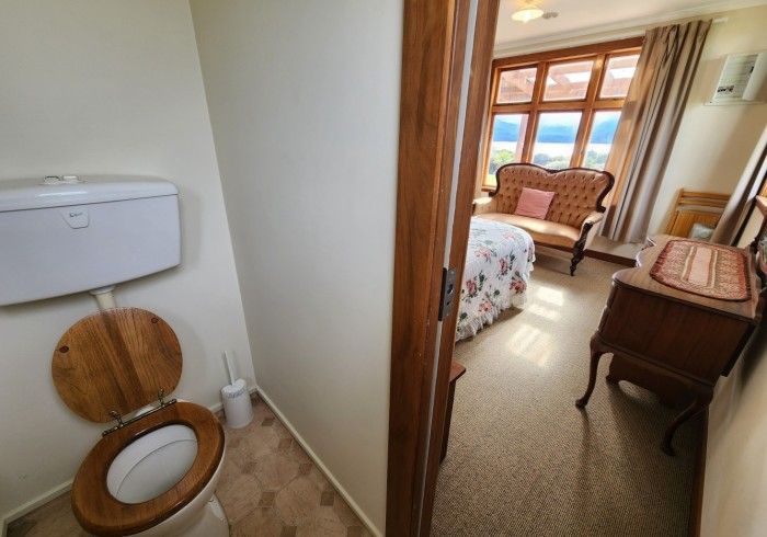 Toilet with wooden seat and tank, doorway view of a bedroom with a small sofa and window.