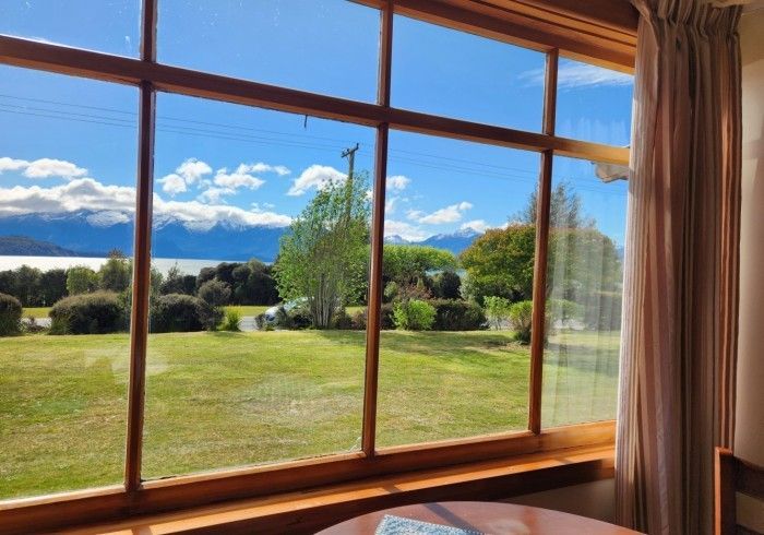 View from a window with a green lawn, trees, and lake against a backdrop of mountains and a blue sky with clouds.