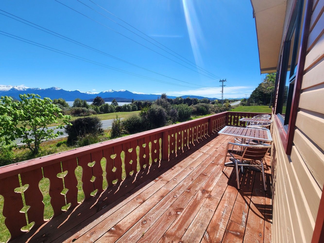 Wooden deck with tables and chairs, overlooking a landscape of fields, water, mountains, and blue sky.