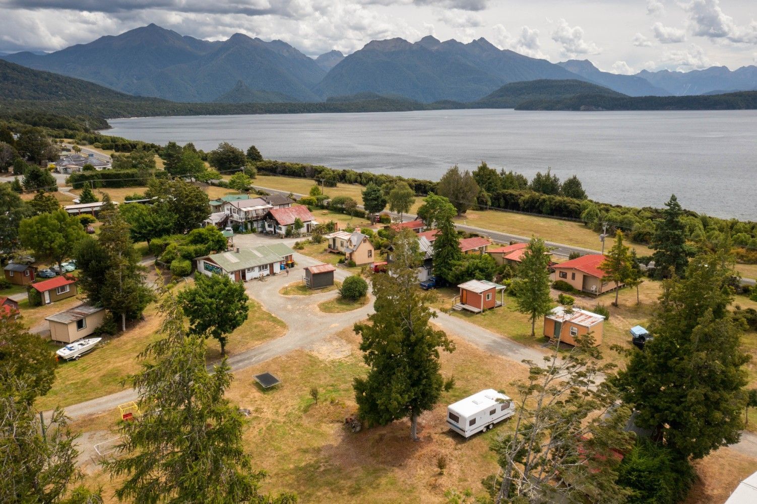 Aerial view of a lakeside town with houses, a campground, and mountains in the background under a cloudy sky.