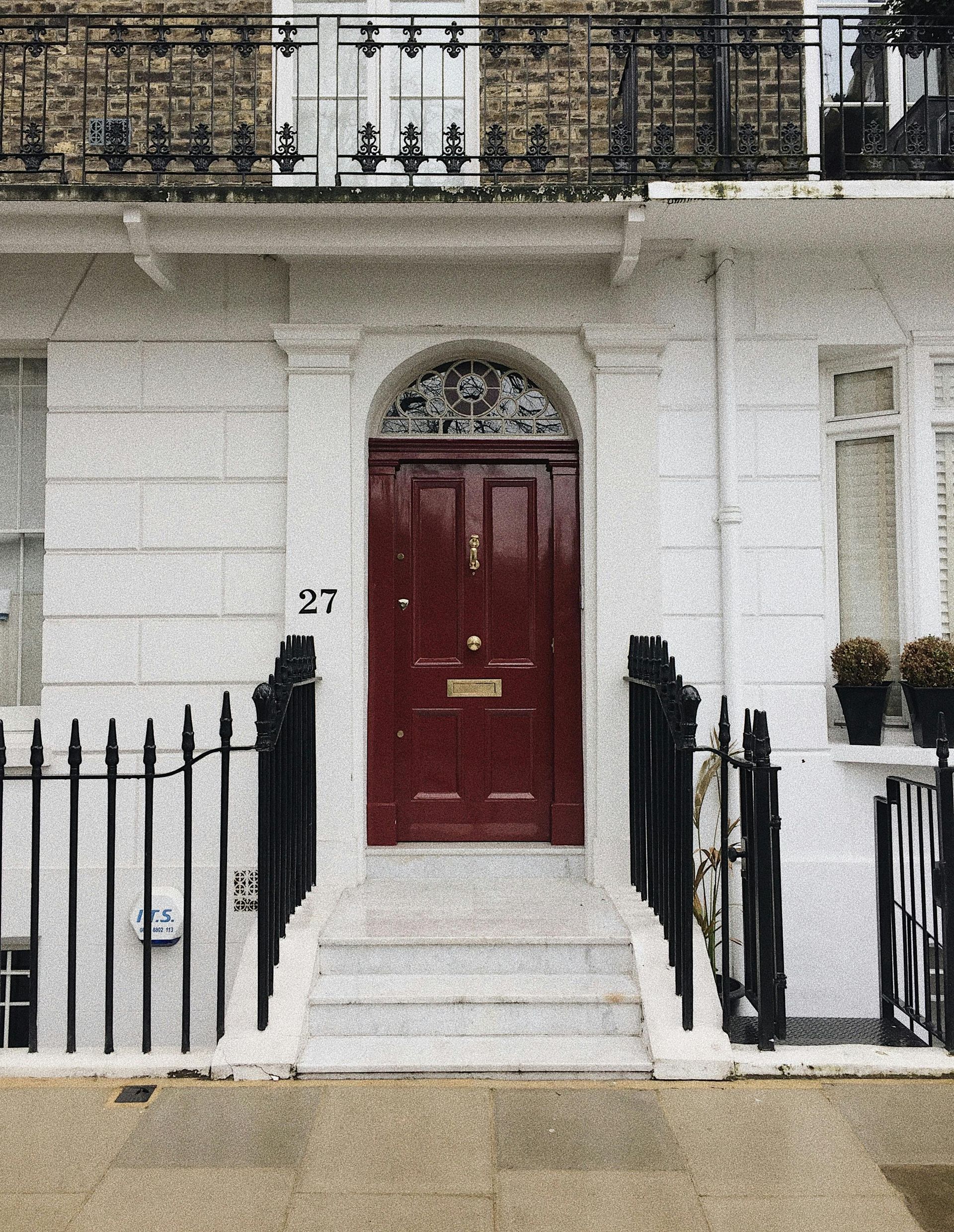 Red door of number 27 framed by white facade, railings, and steps.