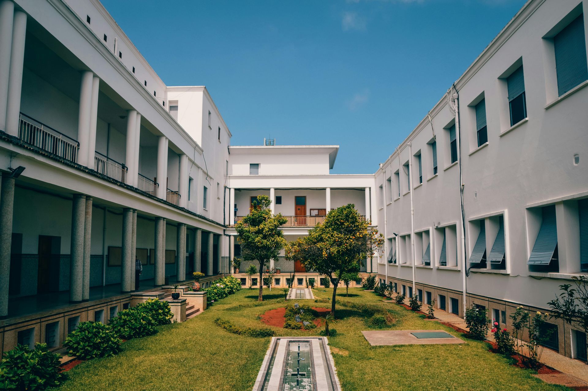 Courtyard with white buildings, green grass, trees, and a fountain under a clear blue sky.