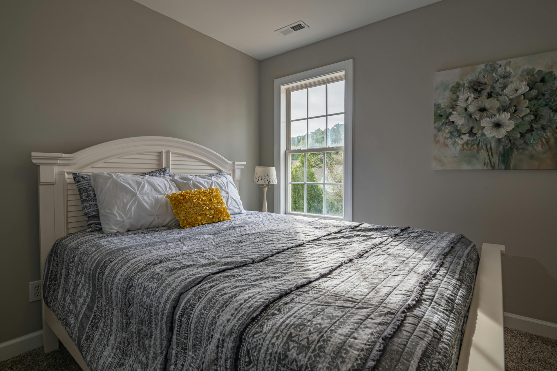 Bedroom with white bed, grey bedding, yellow pillow, window, and floral art.