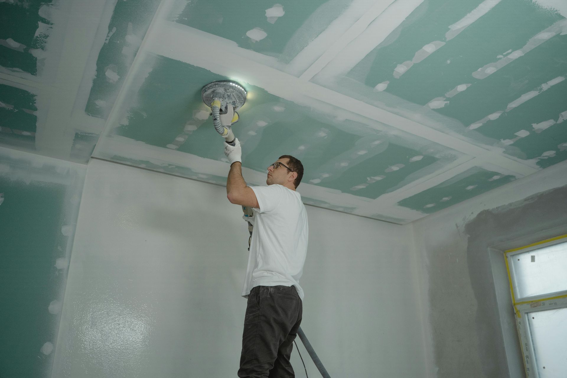 Man sanding a ceiling with a power sander, drywall visible. White shirt, gloves, on a stepladder.