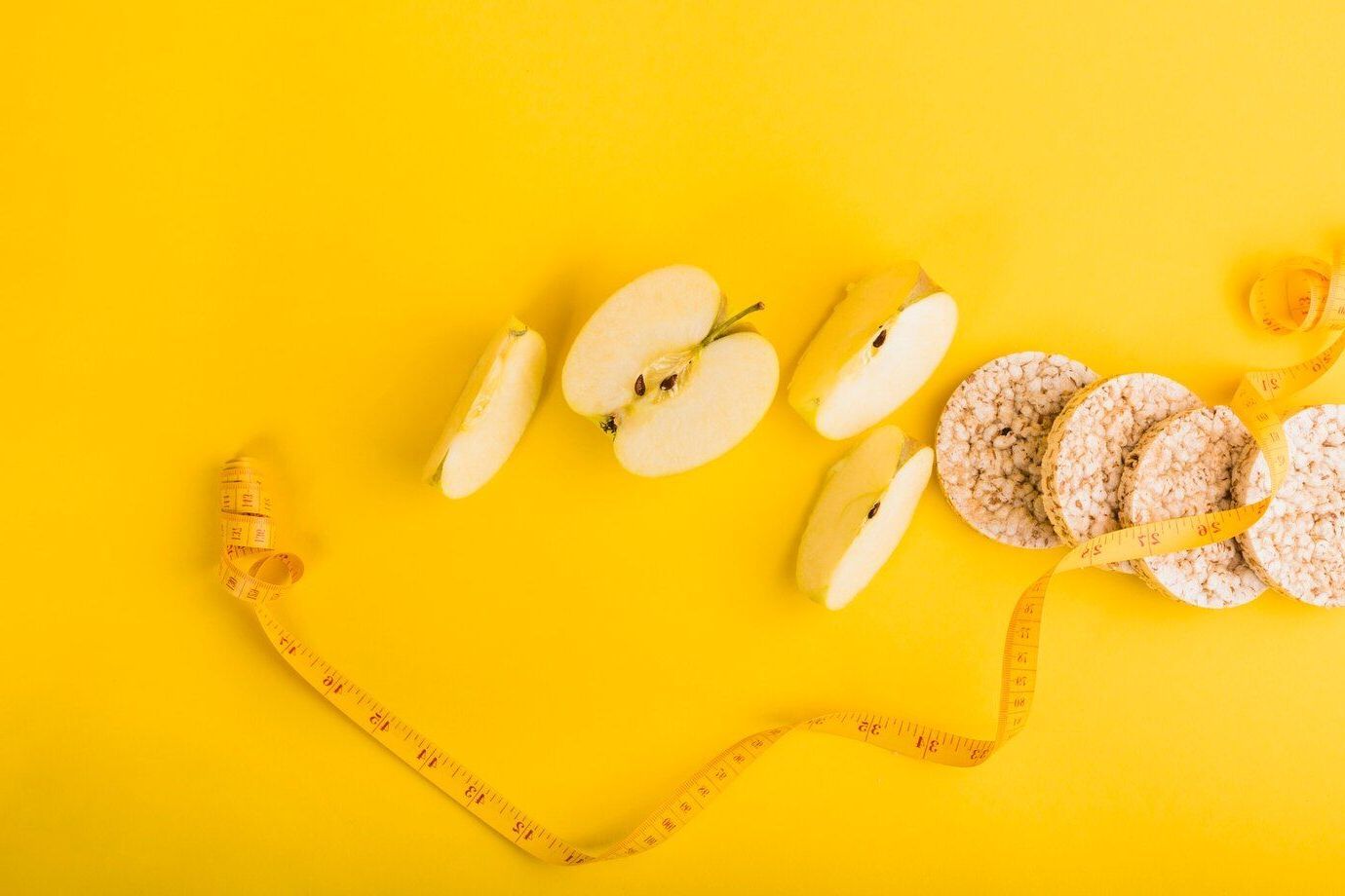 Manzanas, galletas de arroz y una cinta métrica sobre un fondo amarillo.
