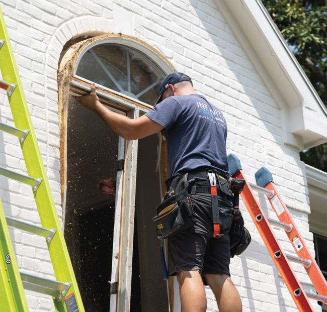 Worker on ladder installing trim around an arched doorway on a white brick house