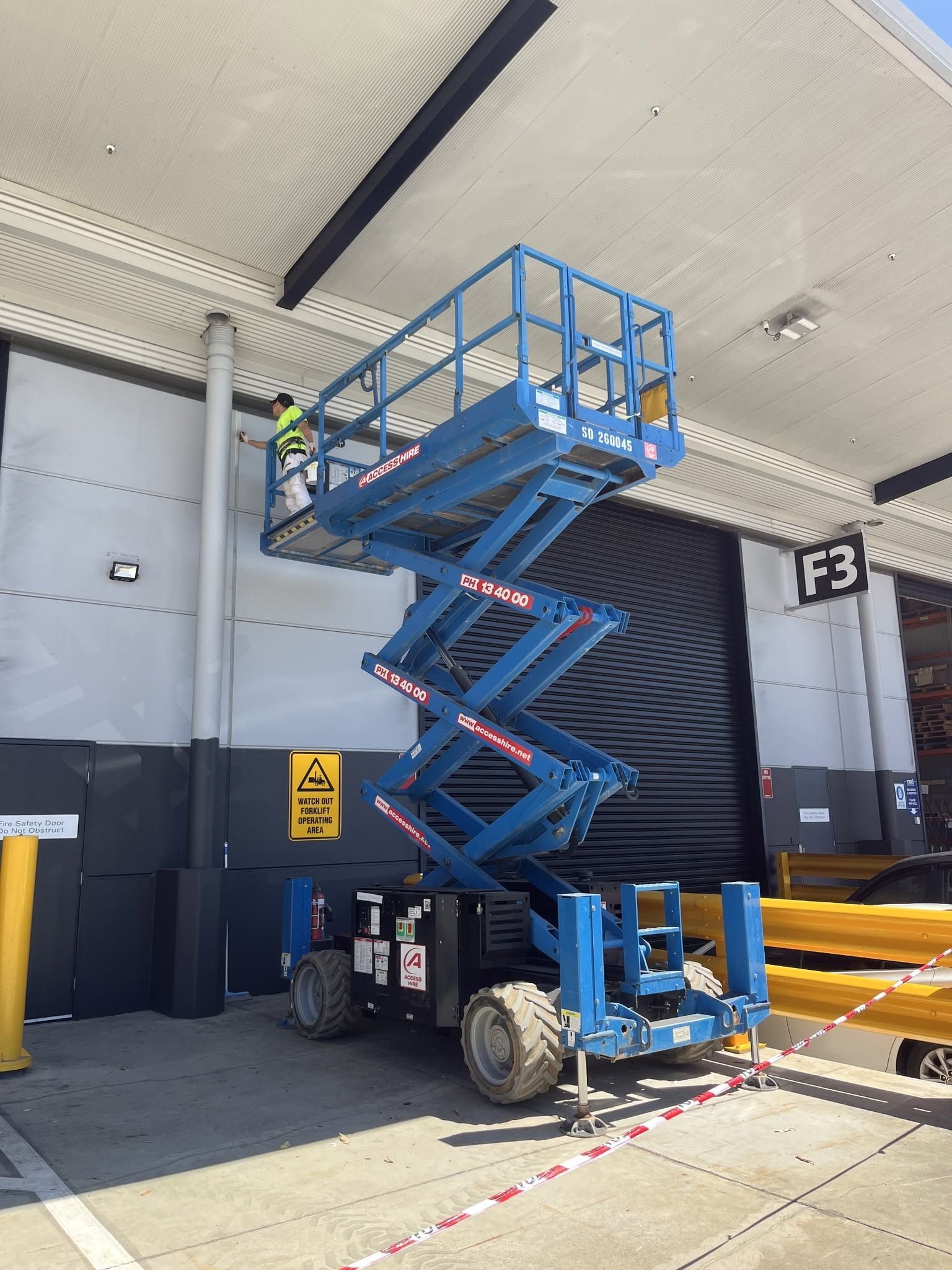 Blue scissor lift raised at a warehouse loading dock under a canopy, with a closed door and yellow safety bollards Blue scissor lift raised at a warehouse loading dock under a canopy, with a closed door and yellow safety bollards