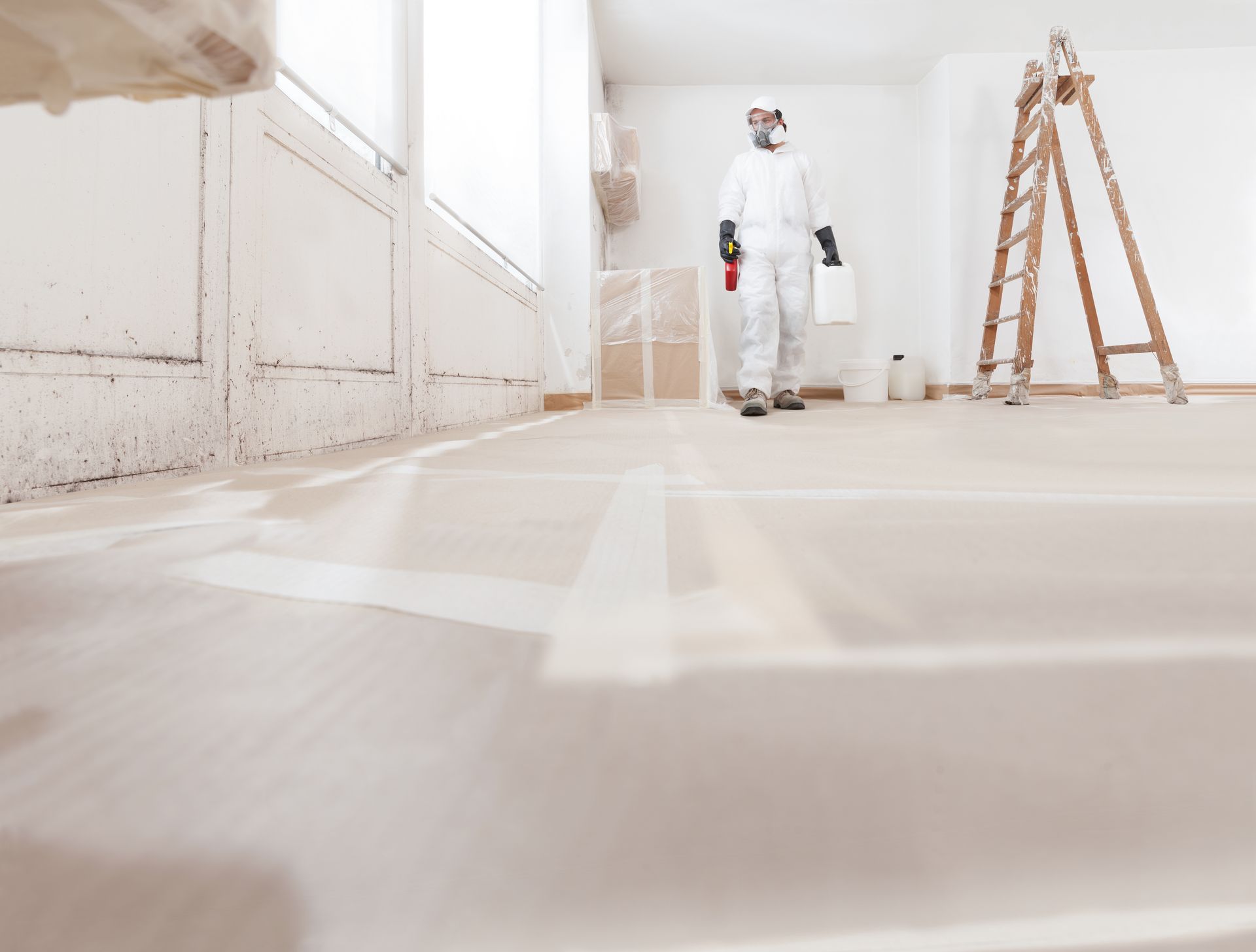 Painter in white coveralls standing in a bright room with a ladder and freshly painted floor lines.