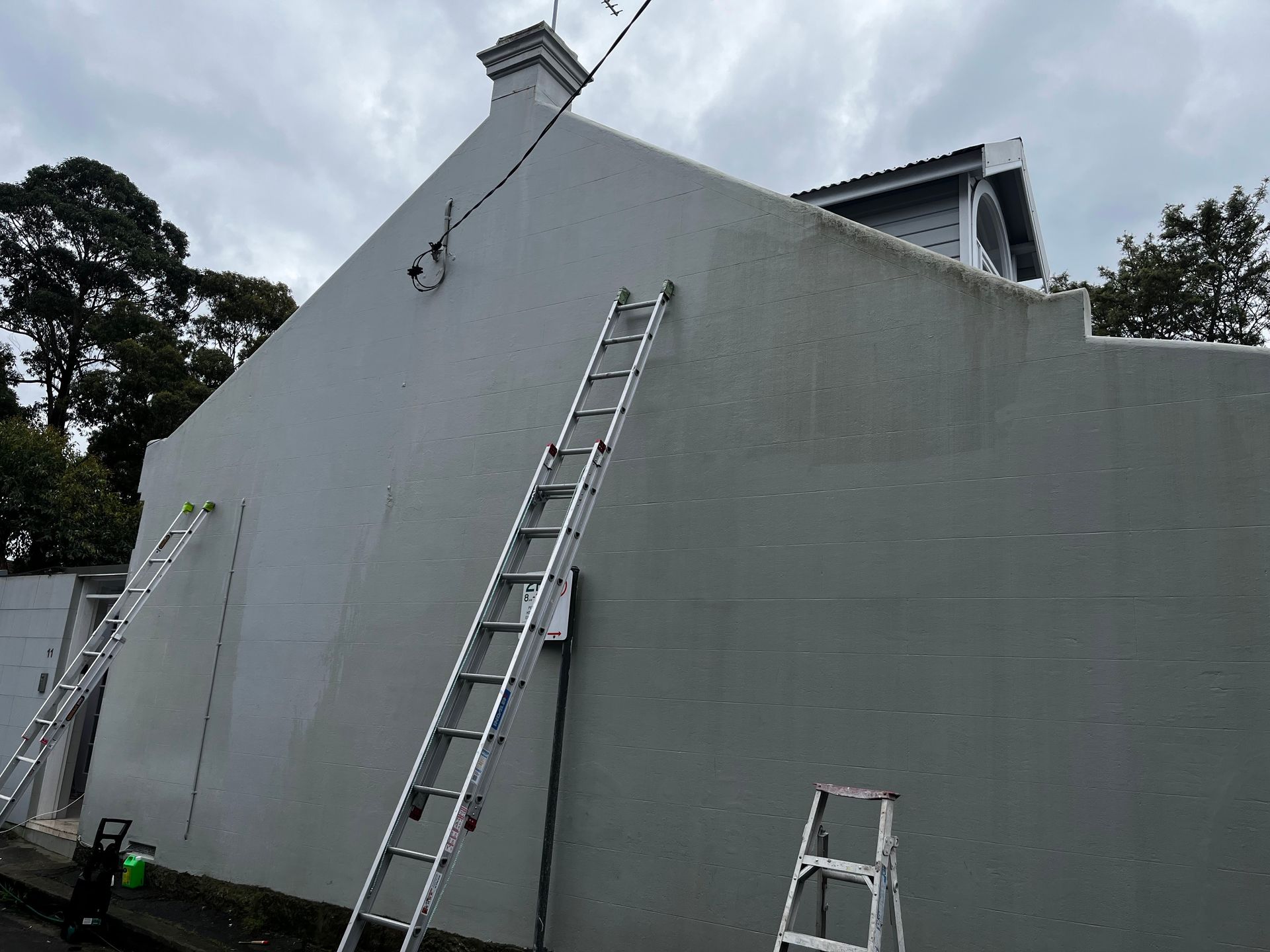 Two metal extension ladders lean against the side of a tall, grey, plastered house wall under a cloudy sky. Two metal extension ladders lean against the side of a tall, grey, plastered house wall under a cloudy sky.