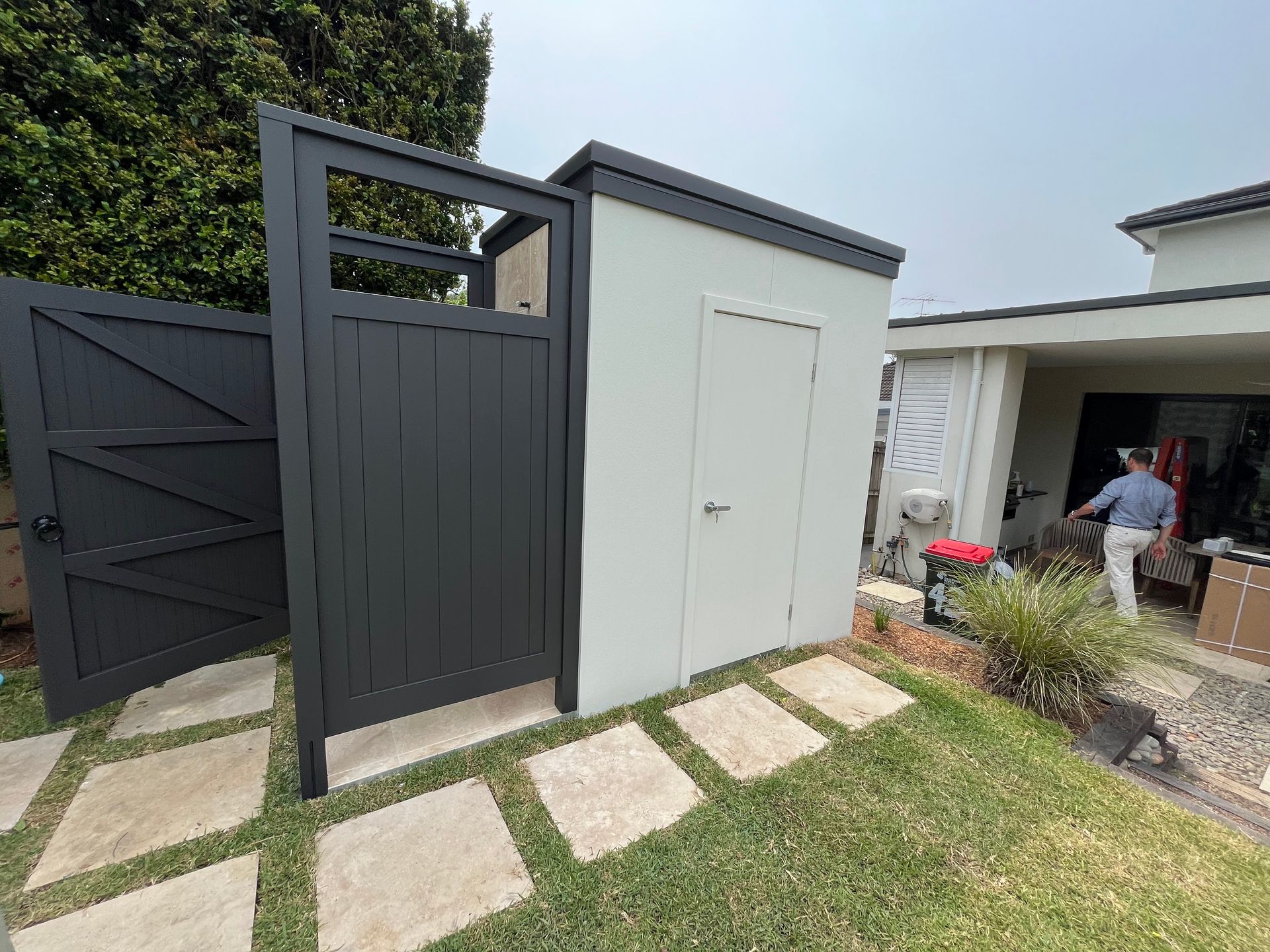A dark charcoal wooden gate and a light gray storage shed in a backyard with stone pavers leading to a house entrance. A dark charcoal wooden gate and a light gray storage shed in a backyard with stone pavers leading to a house entrance.