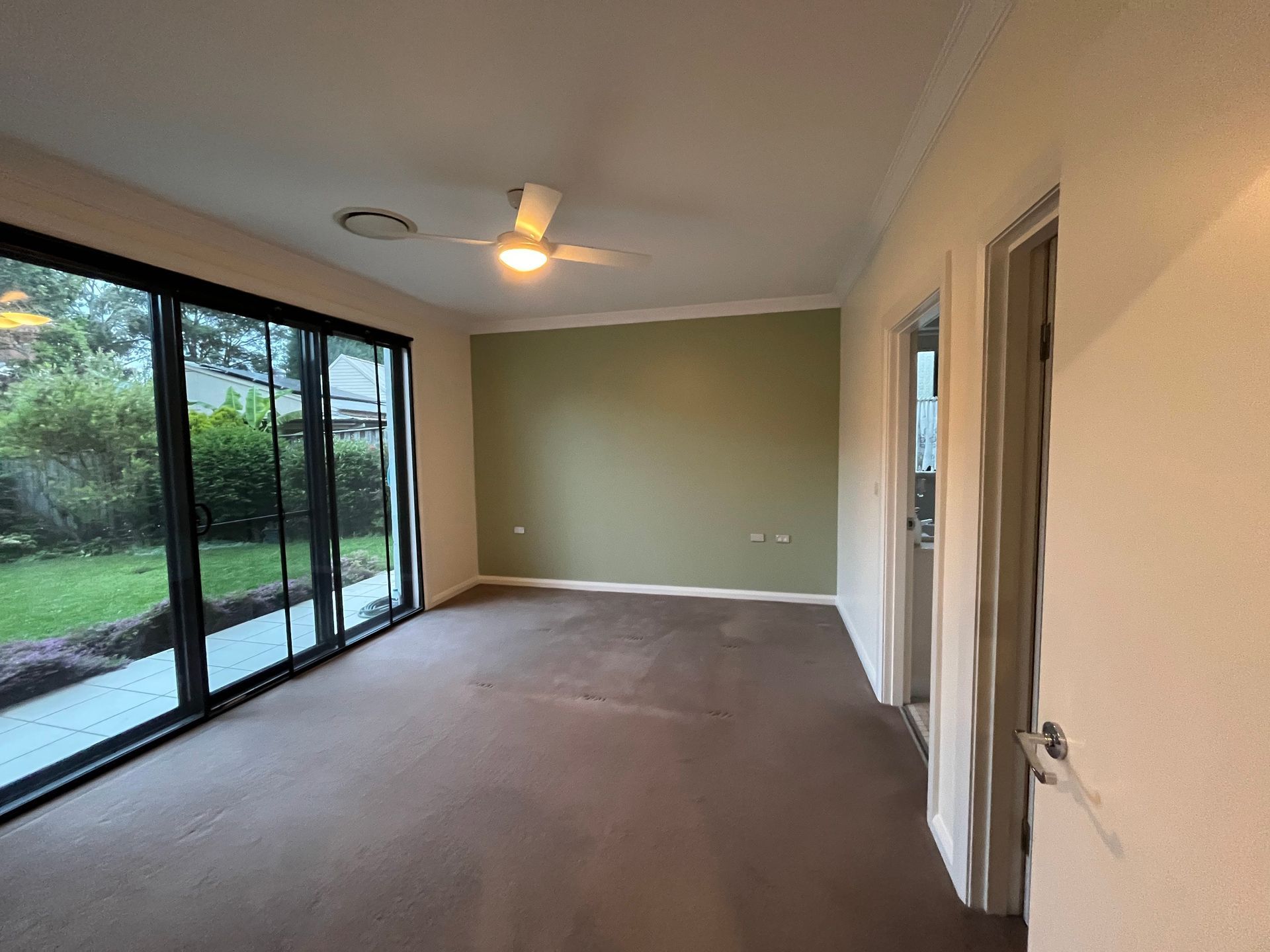 An empty room with tan carpet, a green accent wall, a ceiling fan, and sliding glass doors looking out to a garden. An empty room with tan carpet, a green accent wall, a ceiling fan, and sliding glass doors looking out to a garden.
