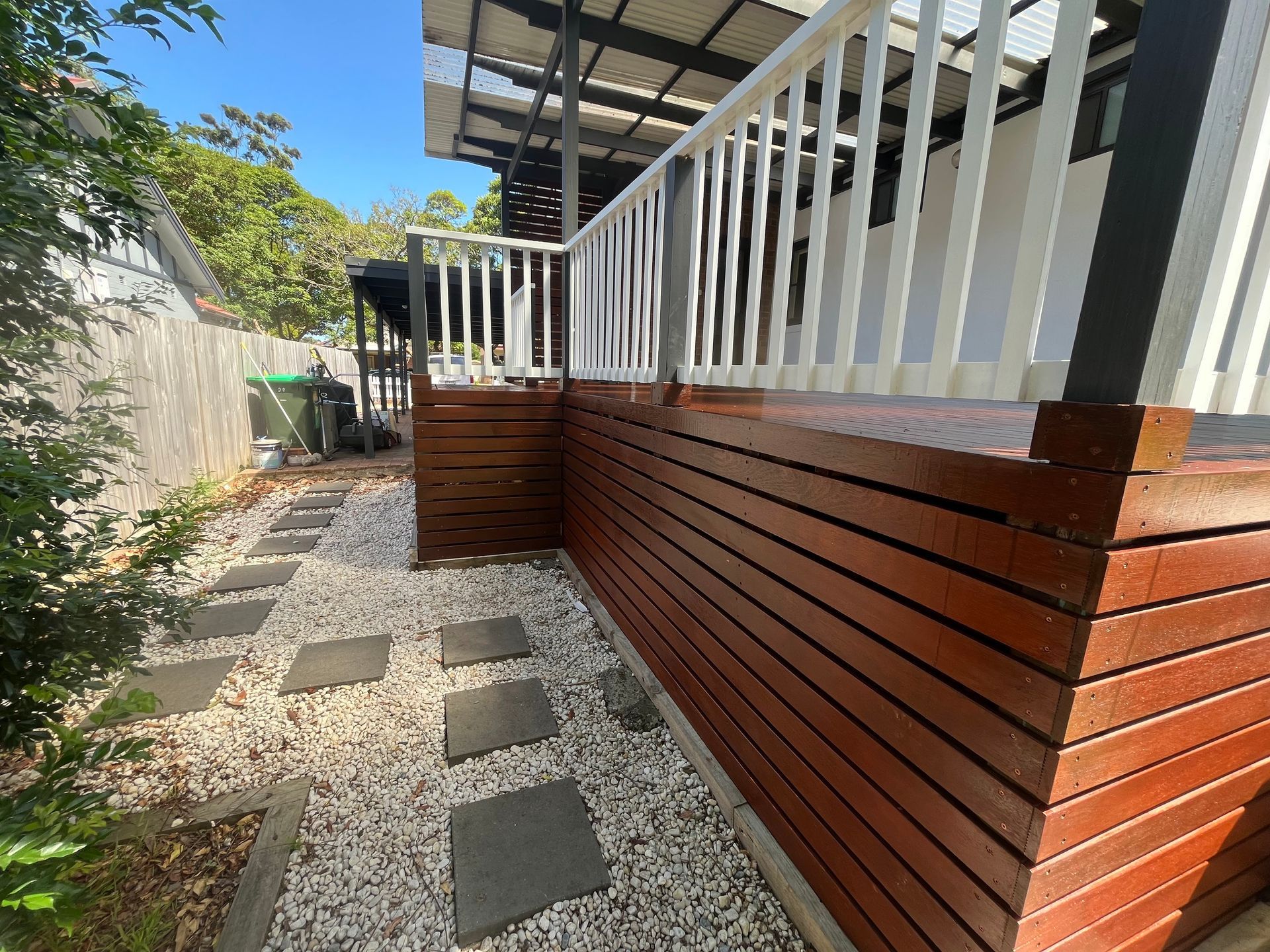 A side view of a raised wooden deck with white railings, set beside a stone path with stepping stones in a backyard. A side view of a raised wooden deck with white railings, set beside a stone path with stepping stones in a backyard.