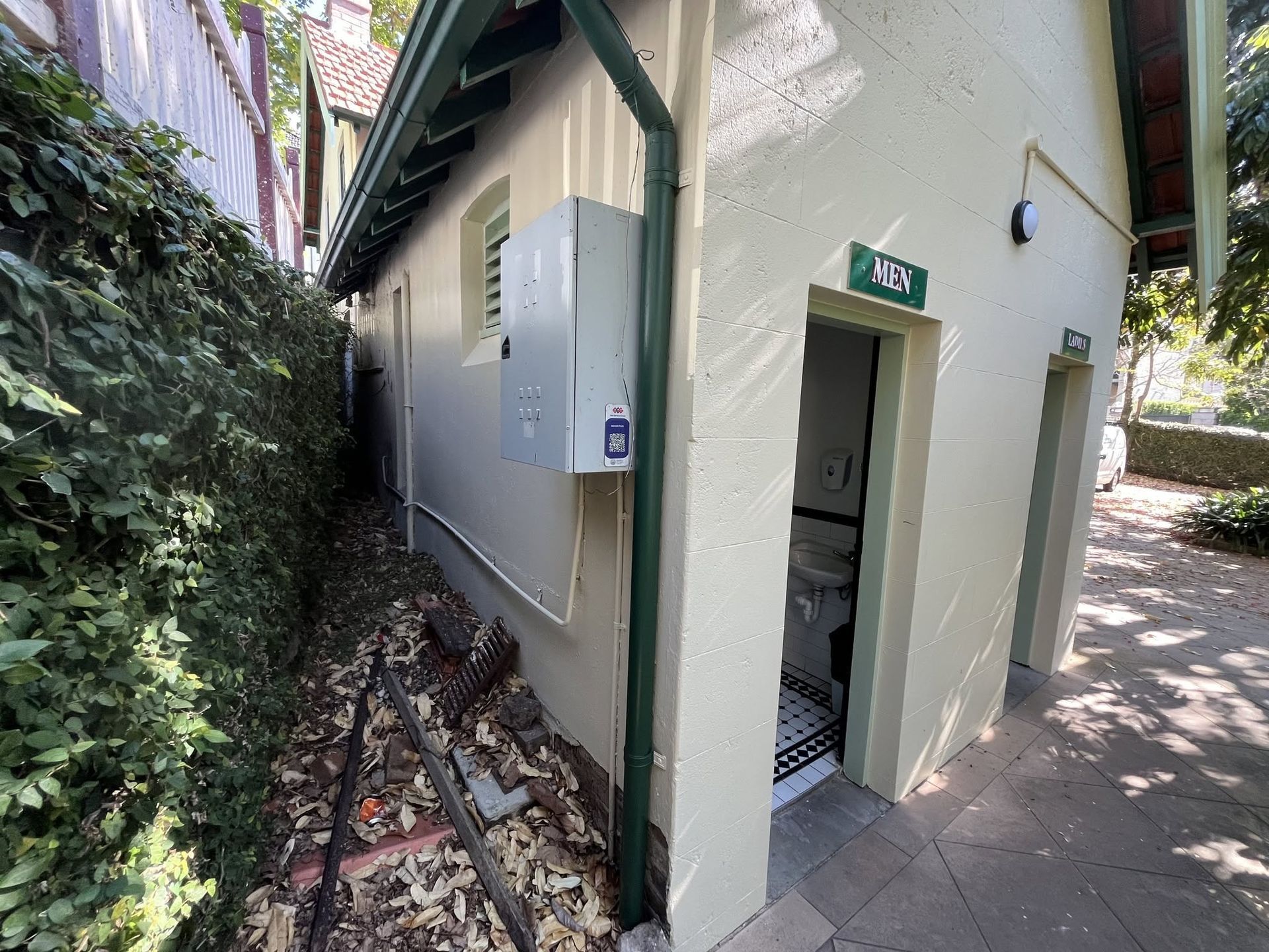 Narrow outdoor walkway beside a small white building with an open door and green trim, next to dense greenery Narrow outdoor walkway beside a small white building with an open door and green trim, next to dense greenery