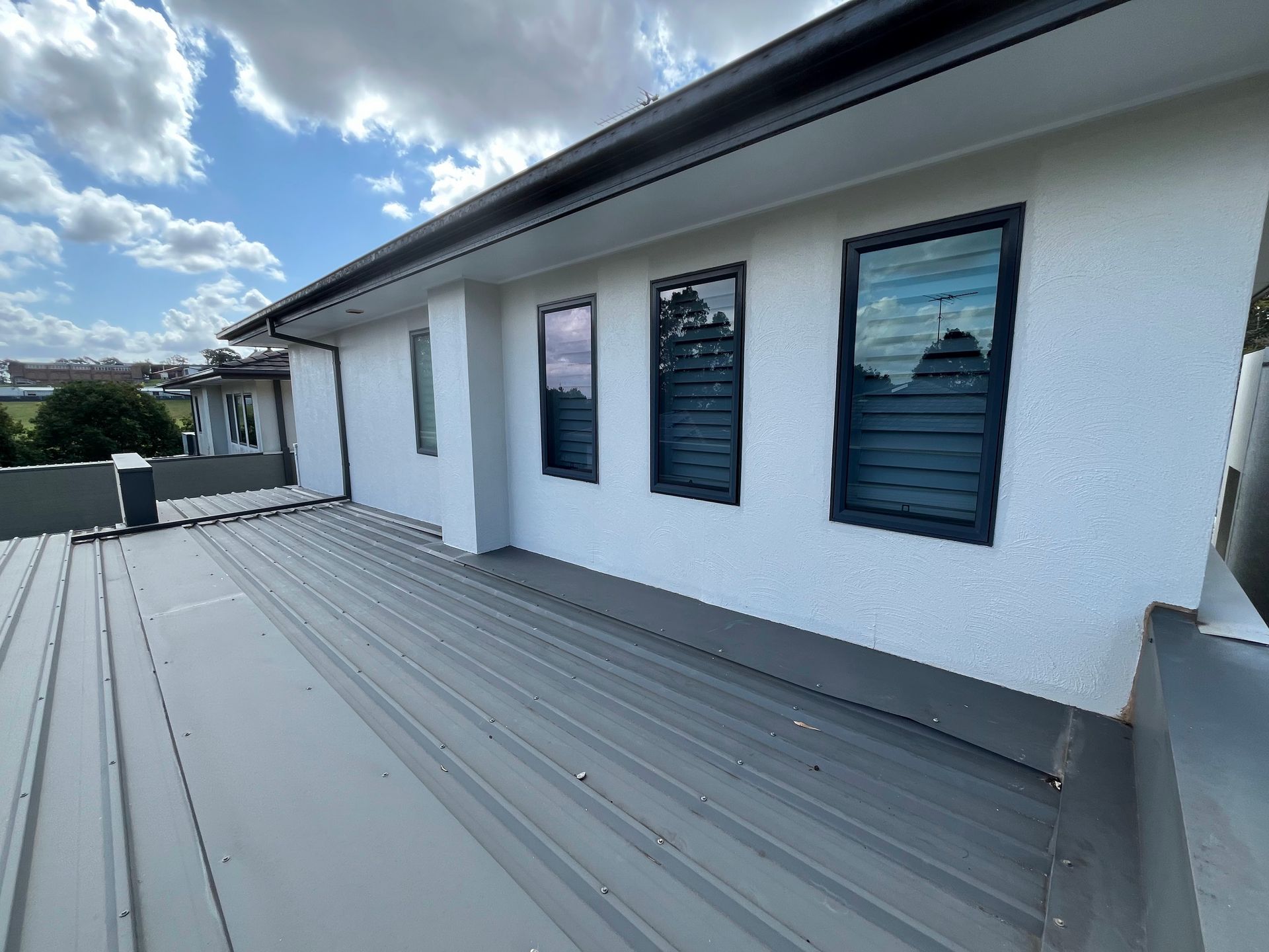 White stucco wall with three dark-framed louvered windows above a gray metal roof under a partly cloudy sky.