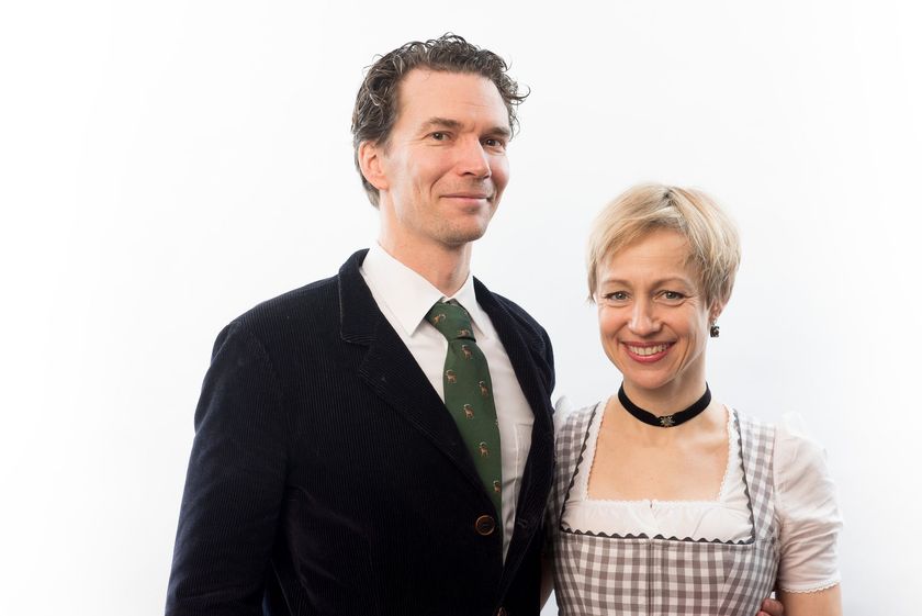 Two people standing against a white background, one in a dark suit and the other in a checkered dress, smiling.