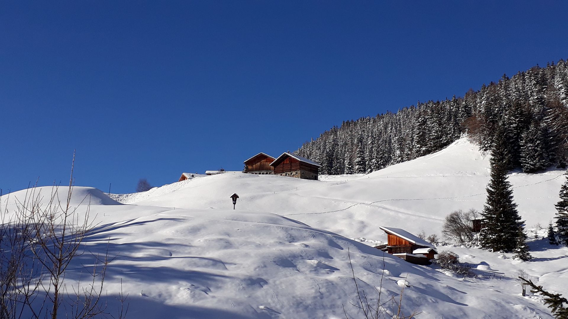 Snowy mountain slope with a person walking towards wooden cabins under a blue sky.