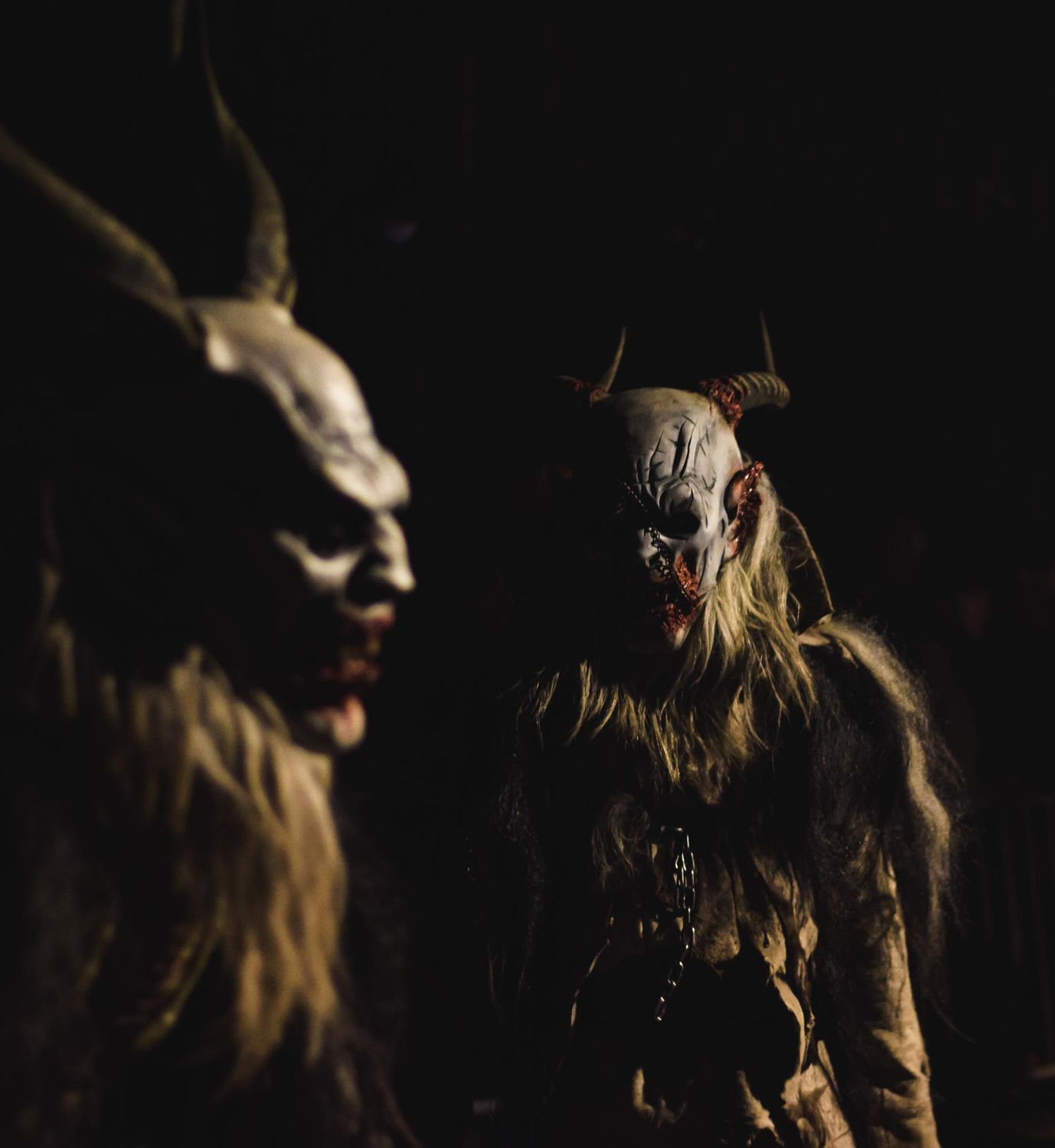 Two Krampus figures with menacing masks, horns, and fur costumes in dark setting.