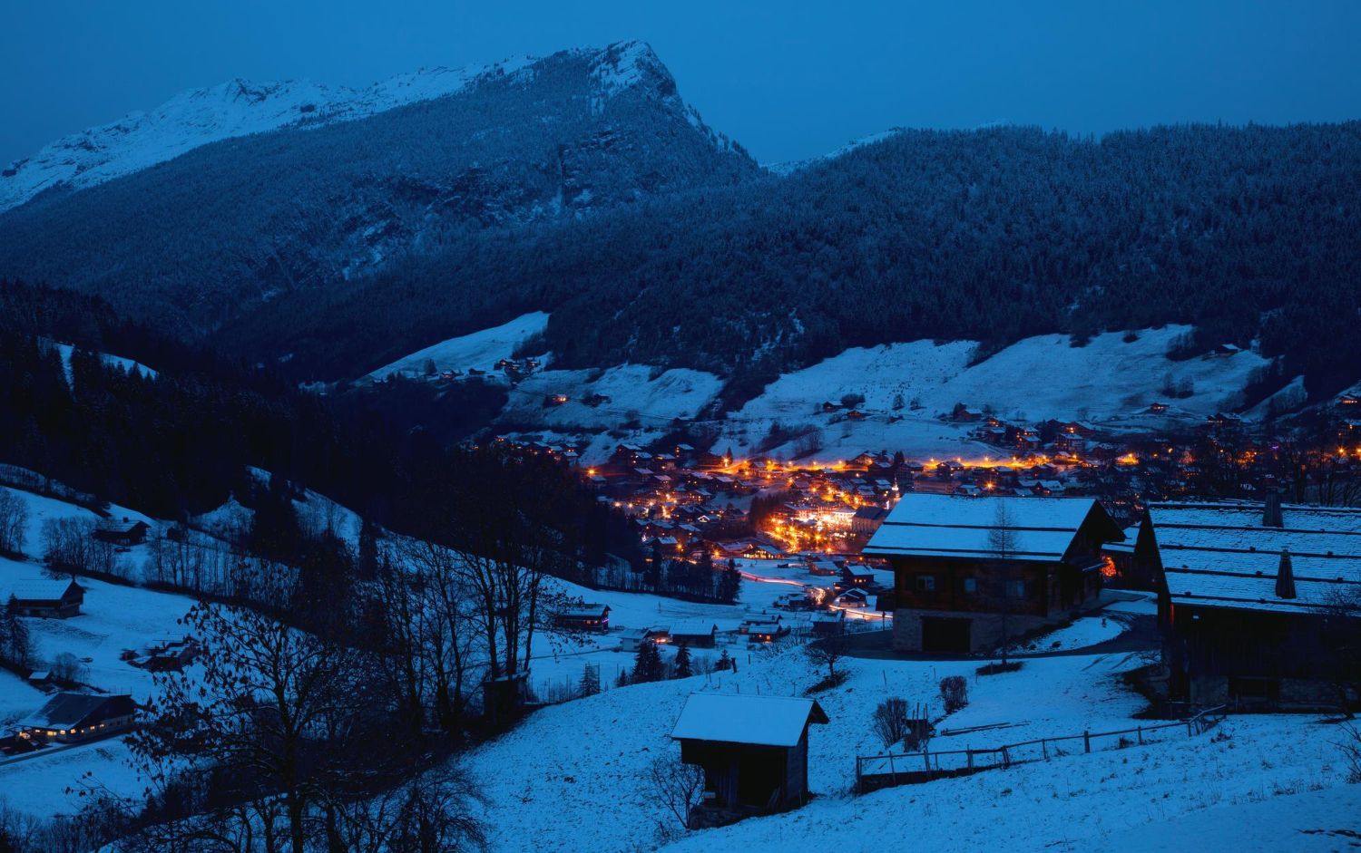 Snowy mountain village at dusk, lit buildings in valley, dark forest and snowy hills in foreground.