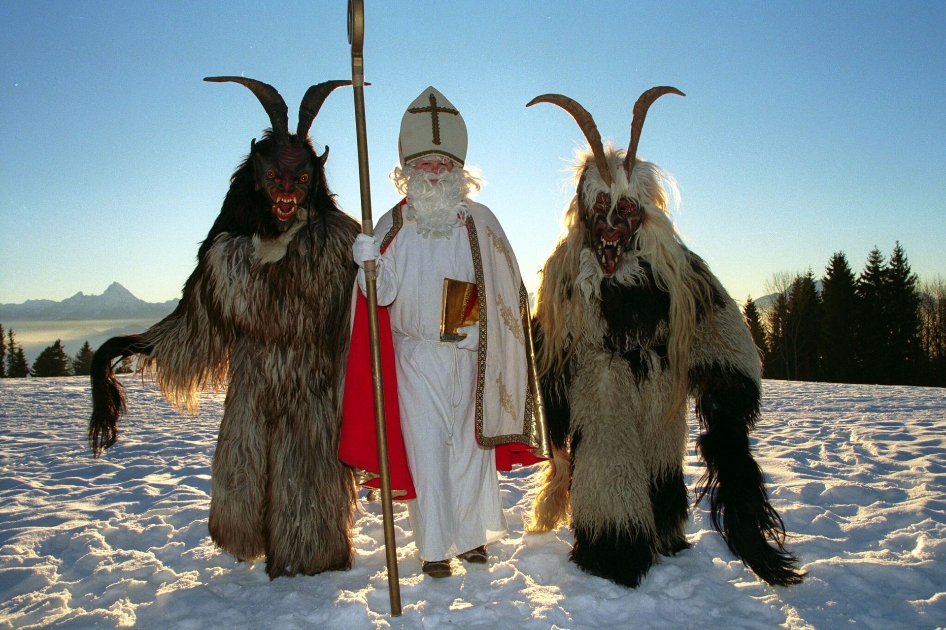Saint Nicholas flanked by two Krampus figures in a snowy outdoor setting.