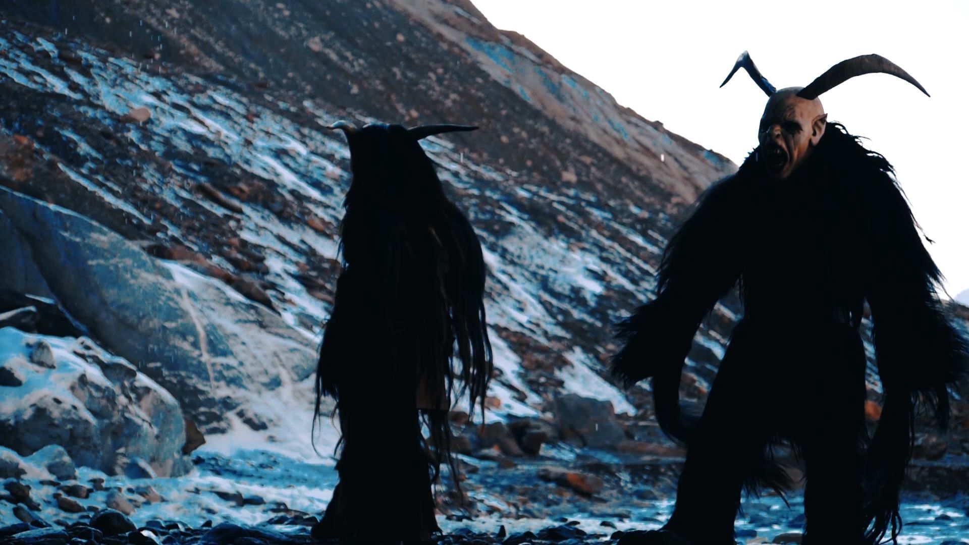 Two figures in dark, furry costumes with horns stand on a snowy mountain.