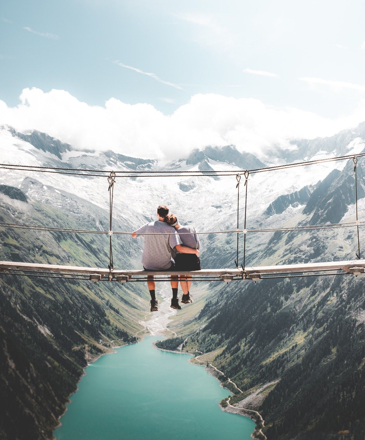 Two people sit on a suspension bridge, looking out over a turquoise lake and snow-capped mountains.