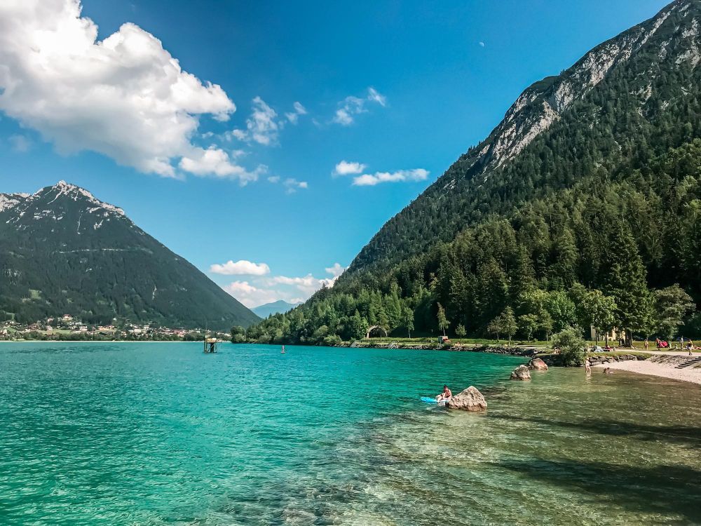 Turquoise mountain lake Lake Achensee with swimmers, forested slopes, and a sandy shore under a blue sky