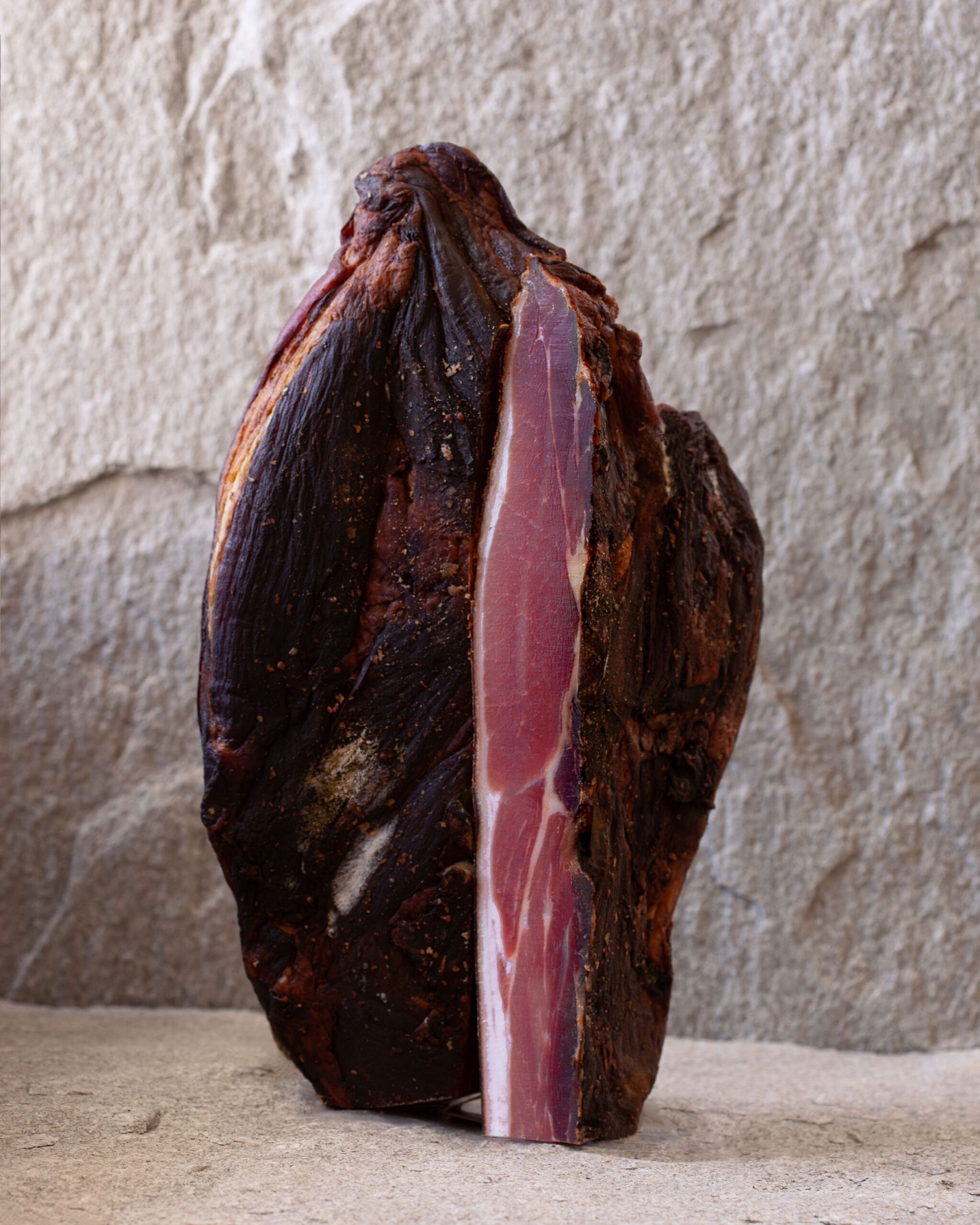 A cured ham, cut to reveal pink interior with a fat streak, against a neutral stone backdrop.