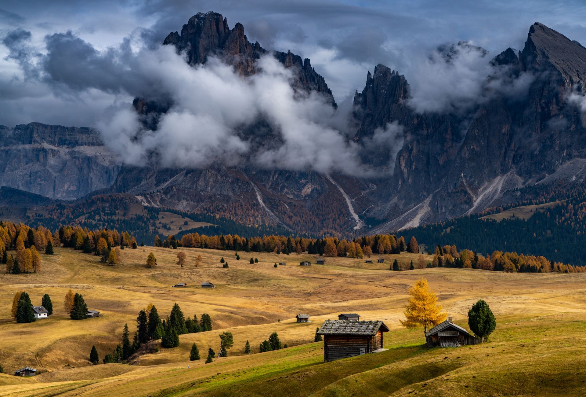 Rolling golden hills with small cabins beneath jagged, cloudy mountain peaks.