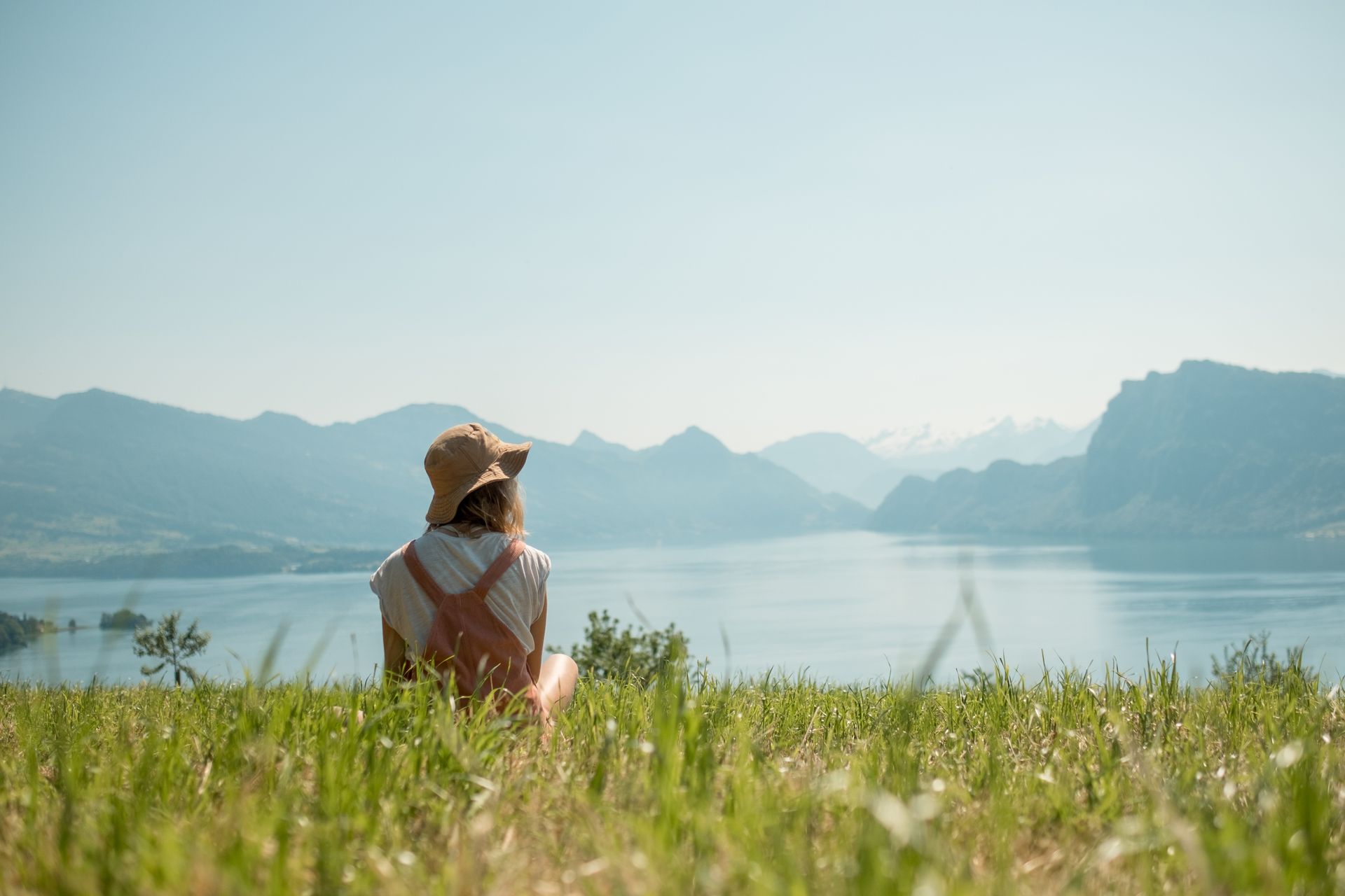 Person in a hat, facing away, sits in tall grass, looking out at a lake and mountains under a bright blue sky.
