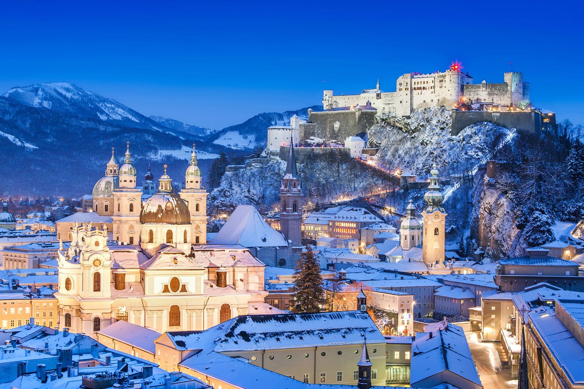 Snowy Salzburg, Austria, featuring illuminated buildings, a fortress on a hill, and snow-covered mountains.