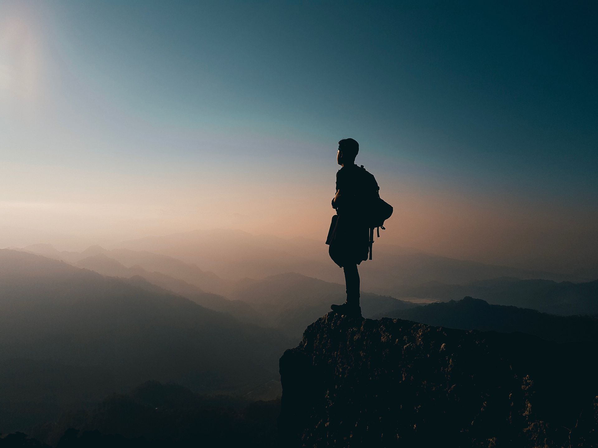 Silhouette of person with backpack standing on cliff, overlooking mountains at sunset.
