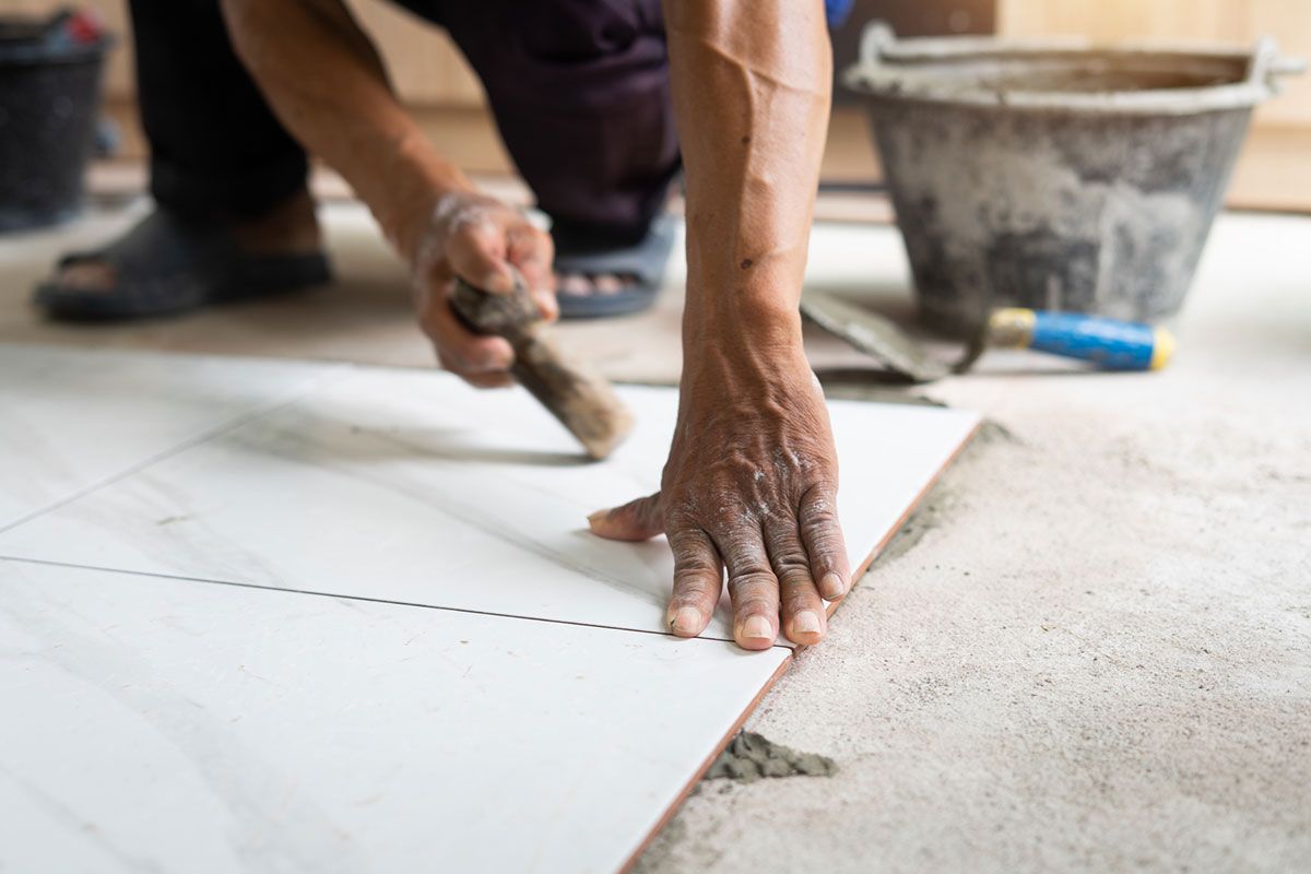 Person tiling a floor, using a trowel. White tile, concrete, bucket in the background. Person tiling a floor, using a trowel. White tile, concrete, bucket in the background.