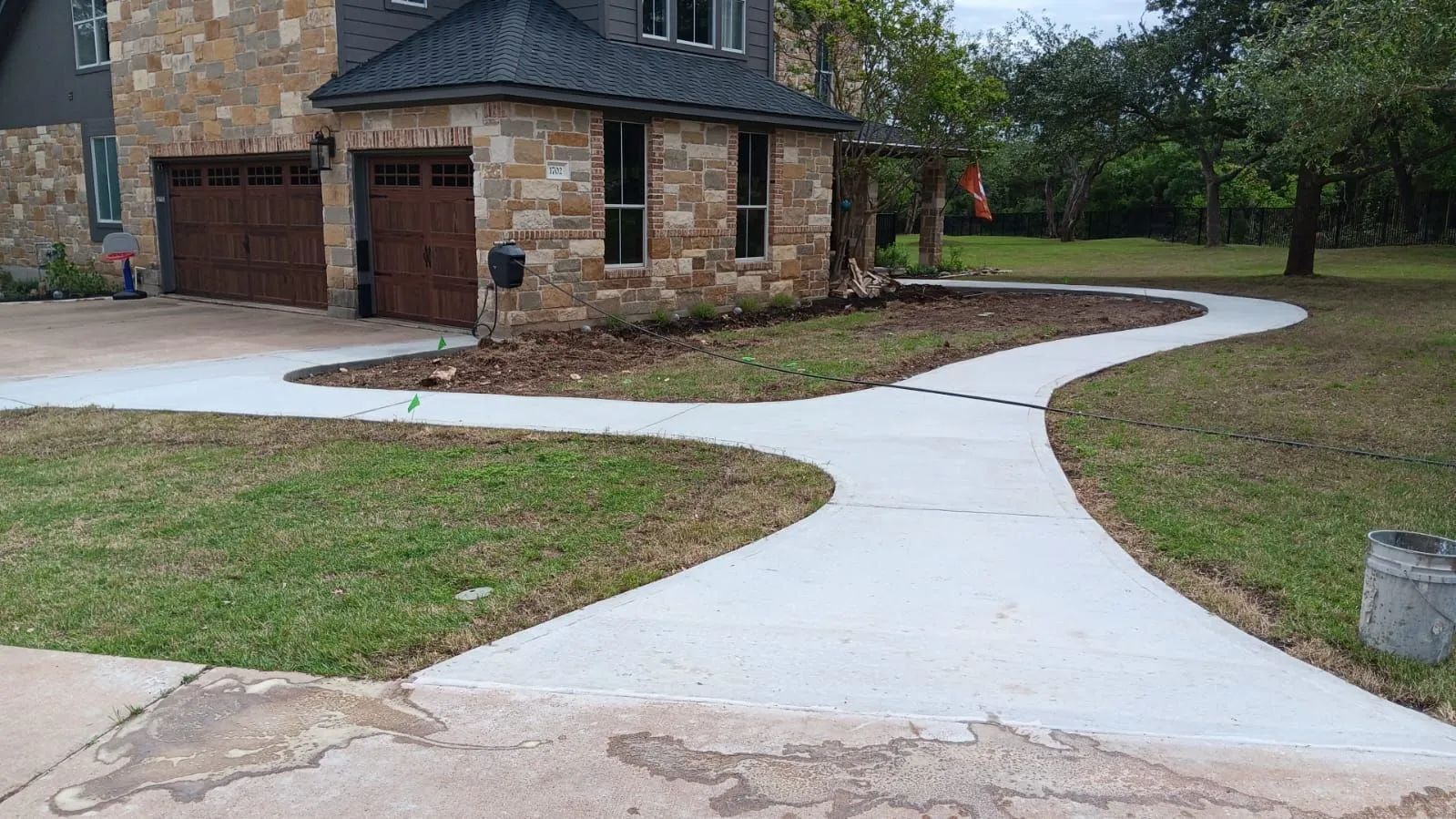 Curved white concrete walkway beside a stone house and landscaped yard with grass and trees