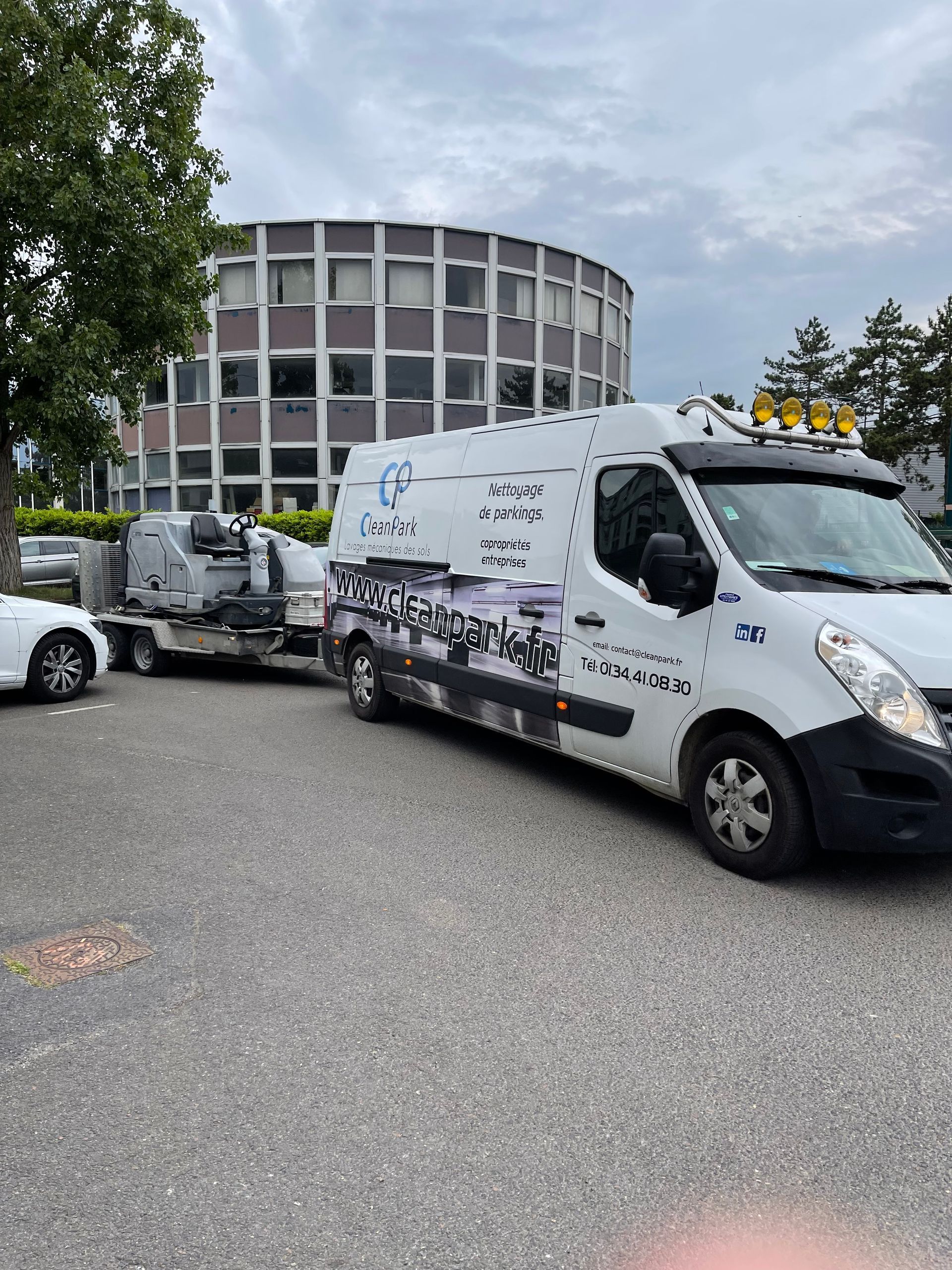 Une camionnette de service blanche est garée devant un bâtiment rond, avec un autre camion derrière, par temps nuageux.